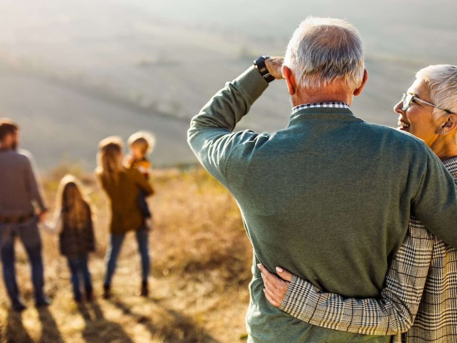 Back view of embraced grandparents enjoying while looking at their family on a hill.