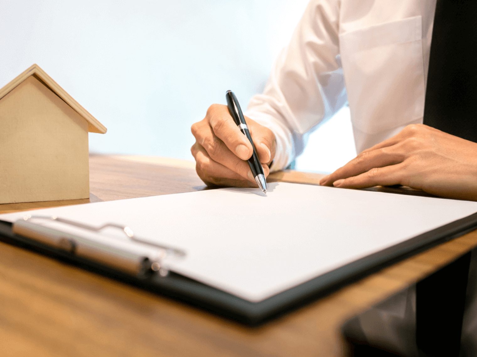 Person in a white shirt and black tie signing a document on a clipboard at a wooden desk with a small house model in the background.