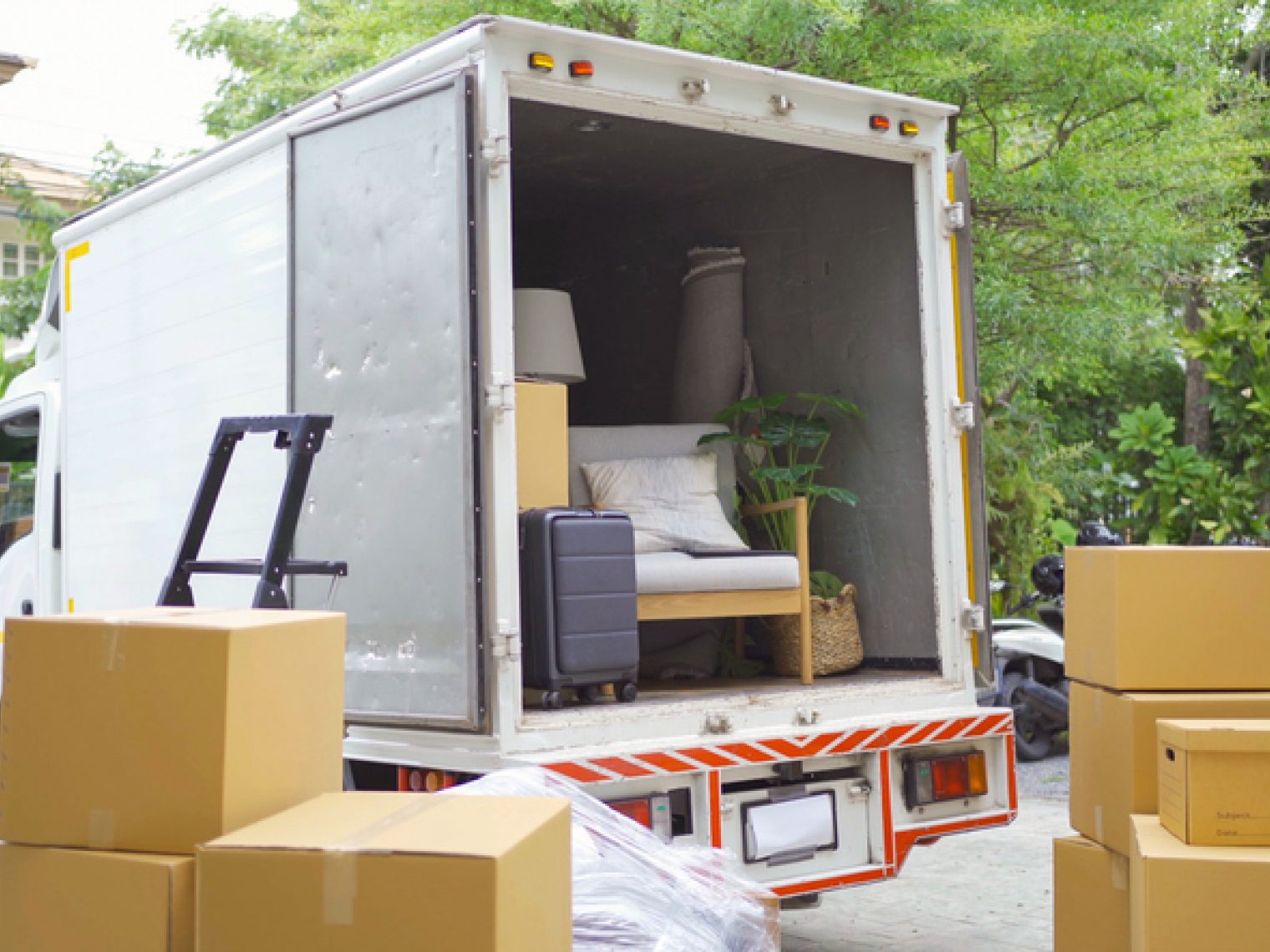 A moving truck is open and partially loaded with household items and furniture, with cardboard boxes stacked in the foreground, indicating a move in progress in a residential area.