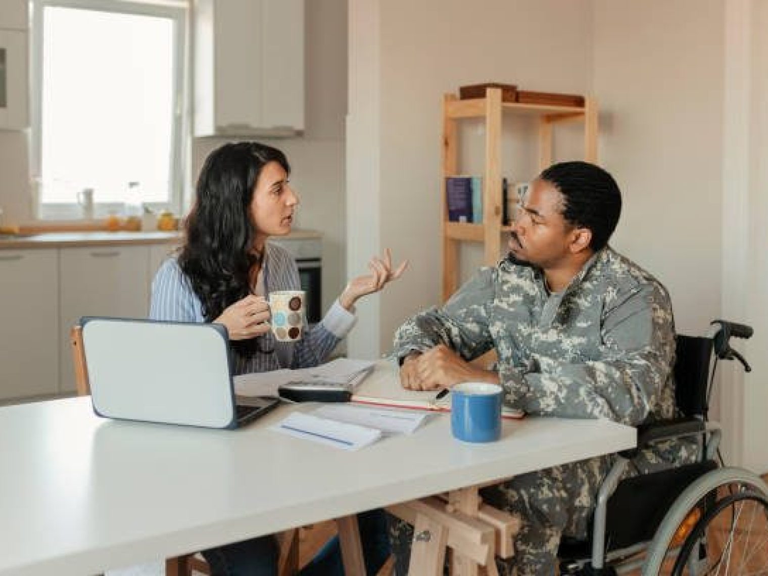 Woman and man in army uniform talking at kitchen table while drinking out of mugs