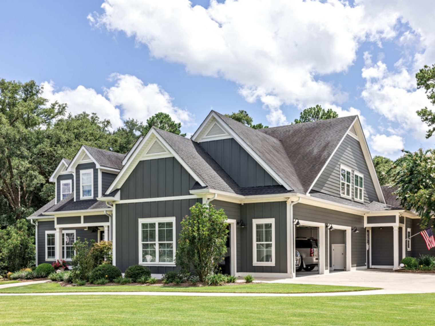 A modern gray house with white trim and an attached garage, set against a backdrop of lush trees and a well-maintained lawn, complete with an American flag displayed prominently.