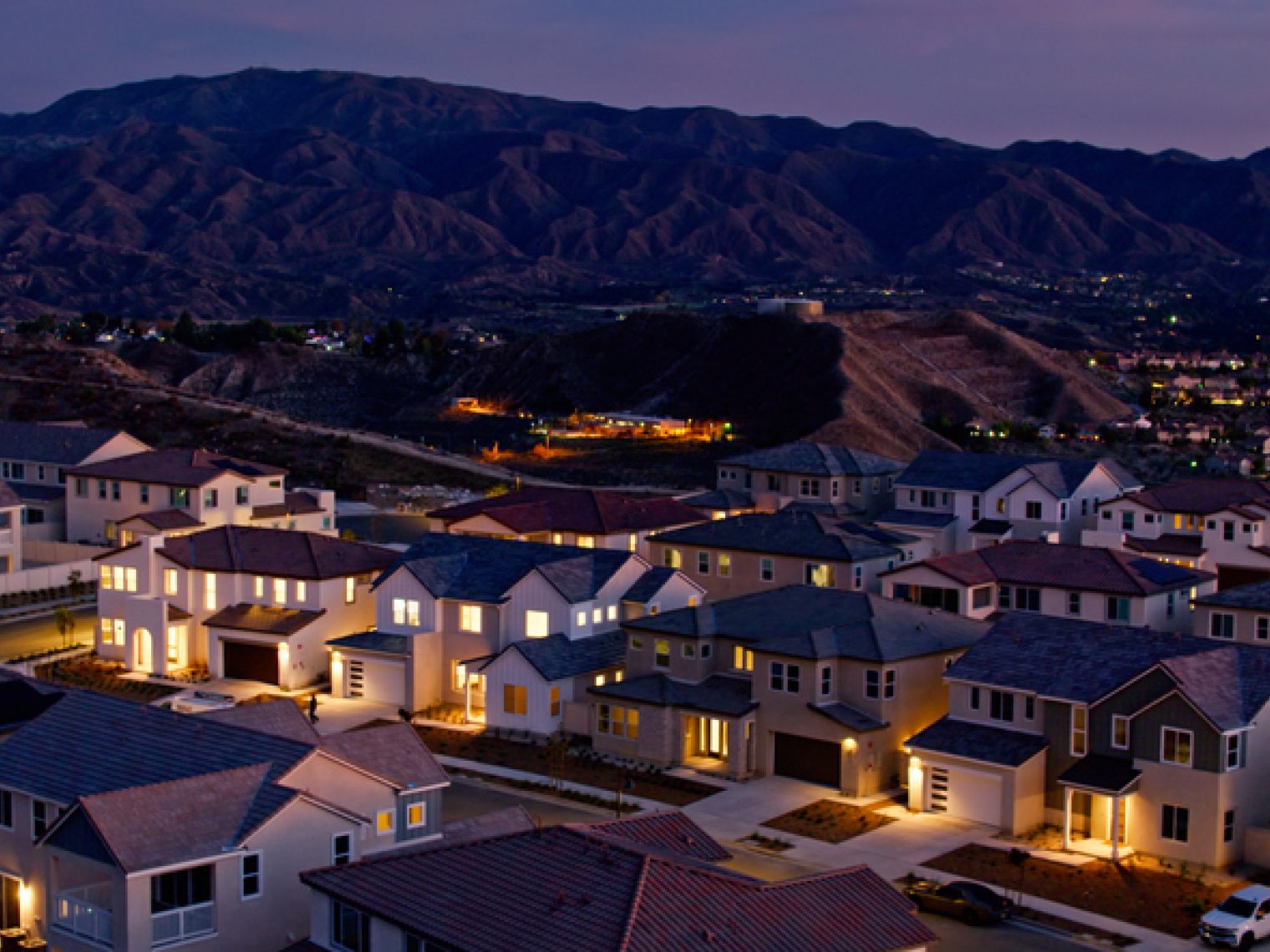 A residential area with modern homes illuminated by evening lights, nestled against a backdrop of rolling hills under a twilight sky.