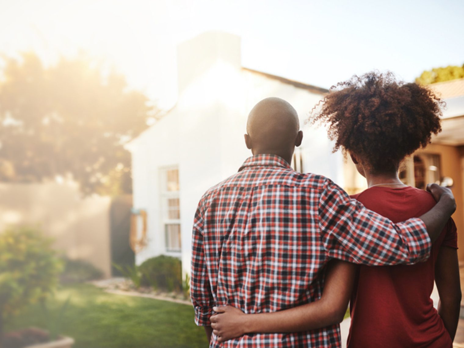 A back view of a couple embracing while looking towards a house with a sunlit garden.