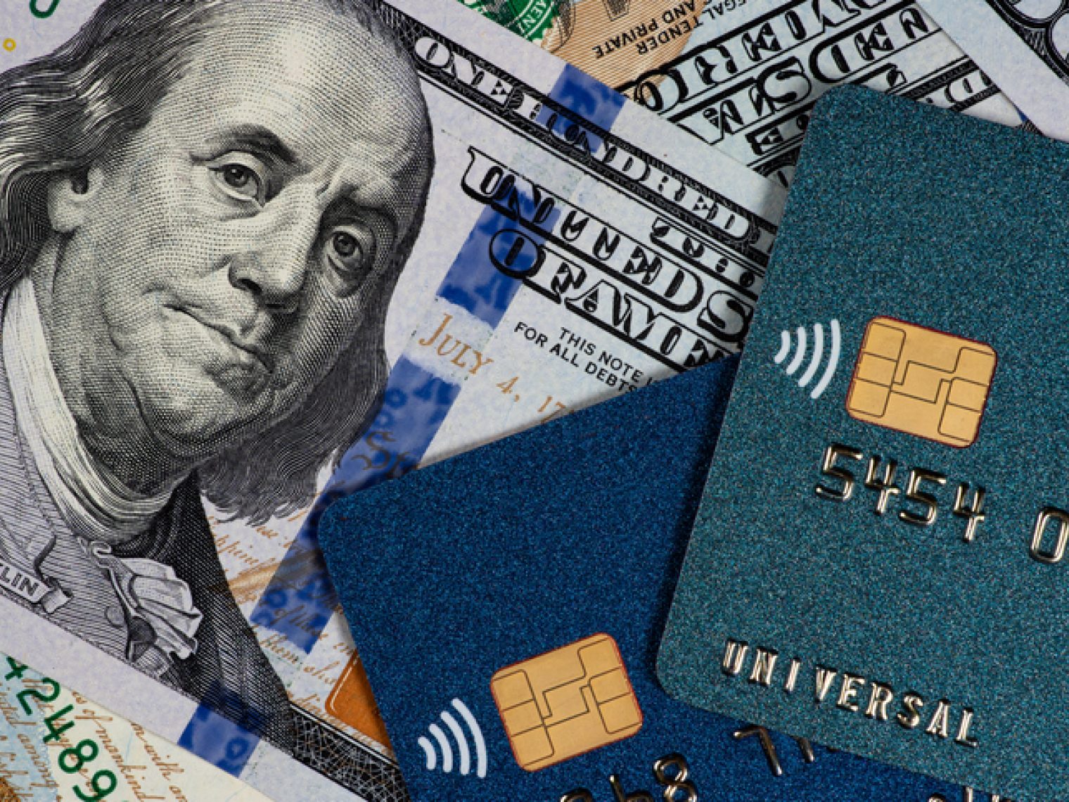 A close-up view of various denominations of U.S. dollar bills with the portrait of Benjamin Franklin visible, overlaid with two credit cards.