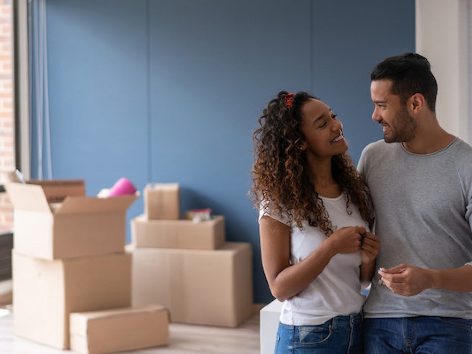 A smiling couple stands close together in a room with unpacked moving boxes, looking affectionately at each other, indicating a new chapter in their lives as homeowners.