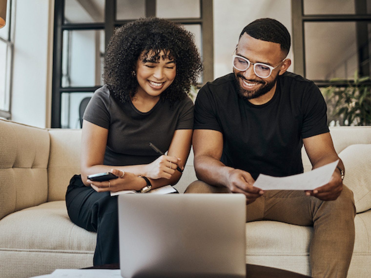 A couple sitting on a couch looking at a laptop smiling with pen and paper in hand.