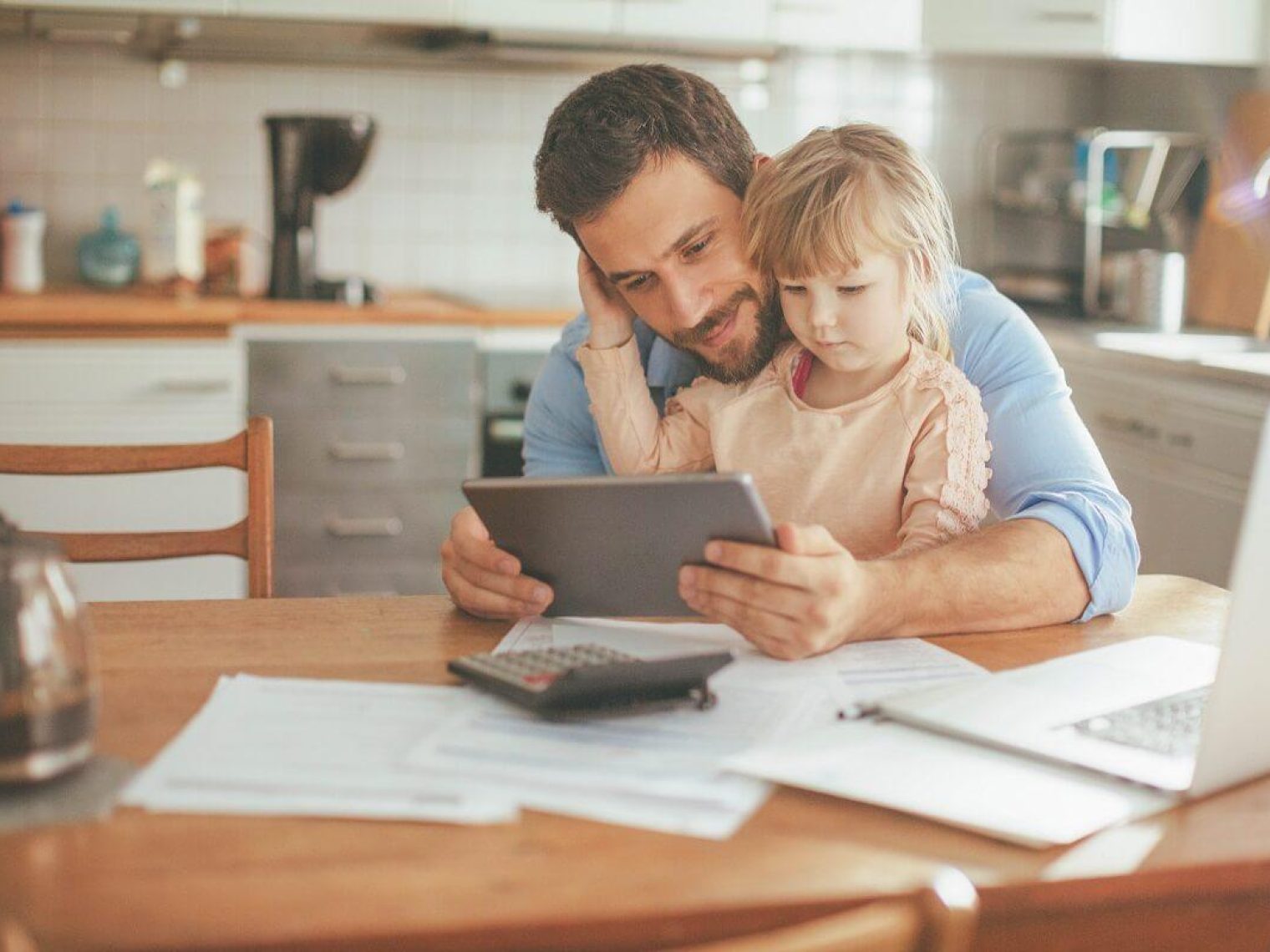 A father and toddler look at a tablet while doing paperwork at the kitchen table.