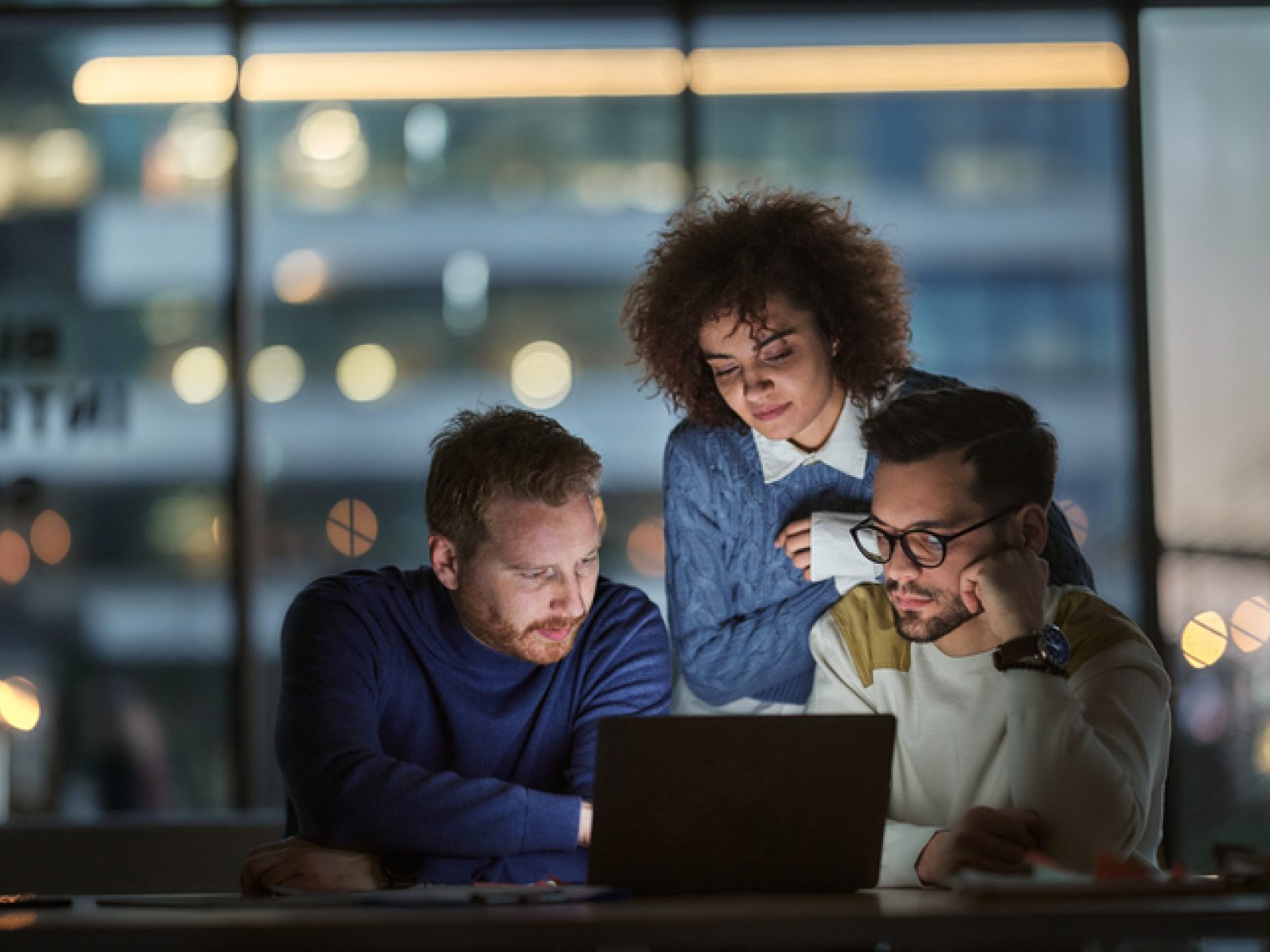 Three colleagues are focused on a laptop screen, working together in an office at night with city lights visible in the background through large windows.