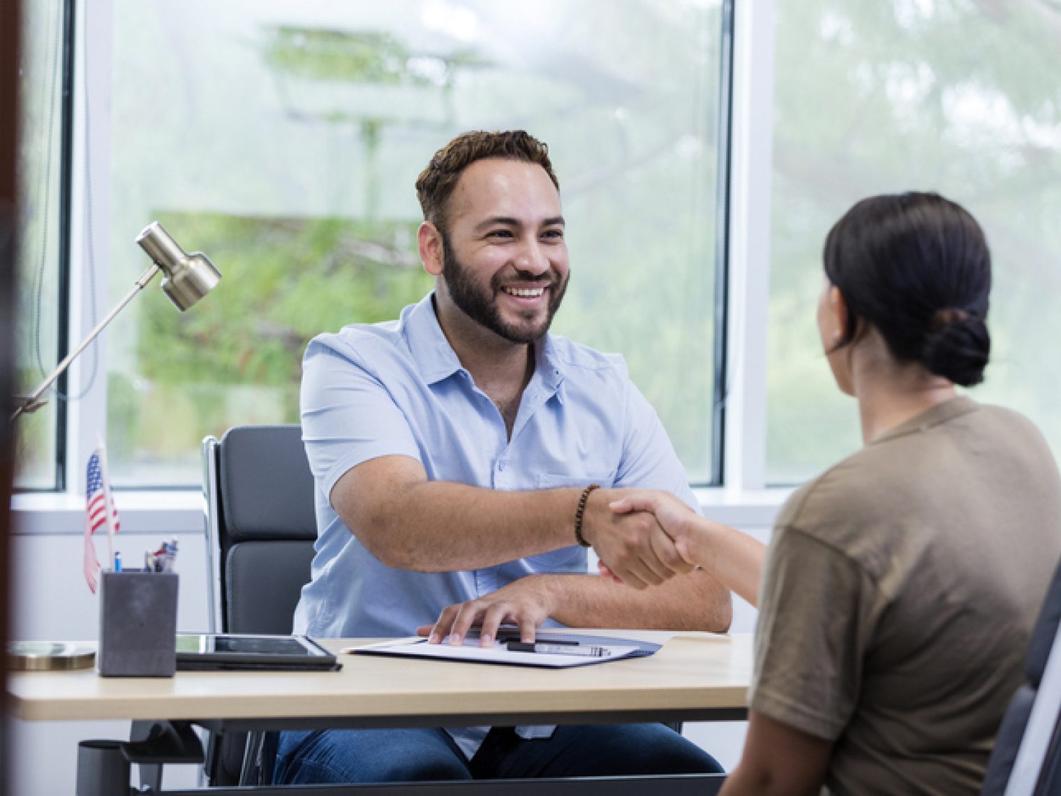 A smiling man extends a handshake across his desk to a woman in a bright office.