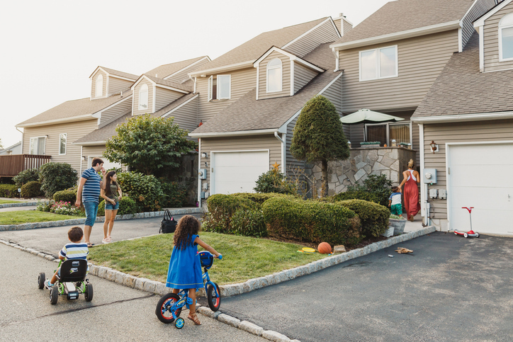 A family is enjoying a sunny day outside their suburban home; two children are playing with their bikes on the driveway, while the adults are in the background by the garage.