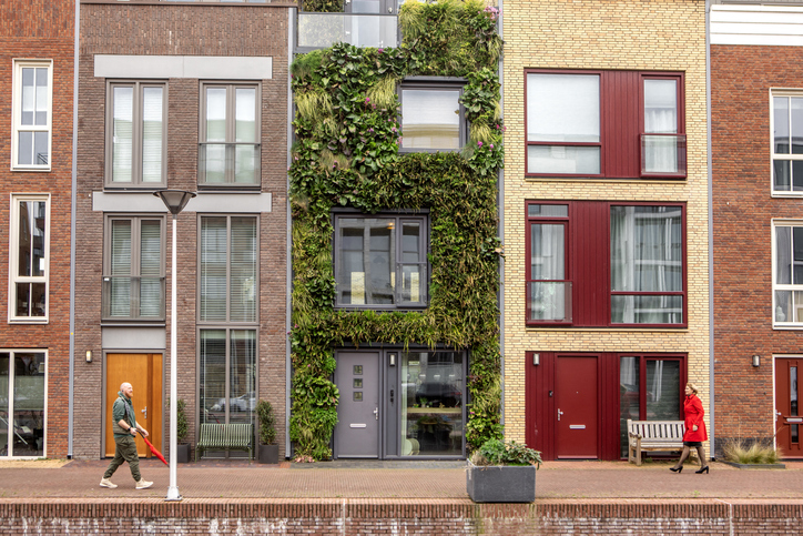 A street view of modern townhouses, one with a vertical garden facade, as pedestrians walk by on the sidewalk, offering a glimpse into urban residential architecture.