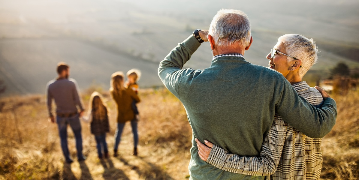 Back view of embraced grandparents enjoying while looking at their family on a hill.