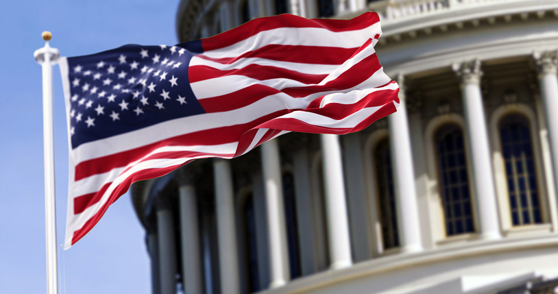 The United States flag flies prominently in front of a classical government building with iconic columns, against a clear blue sky.