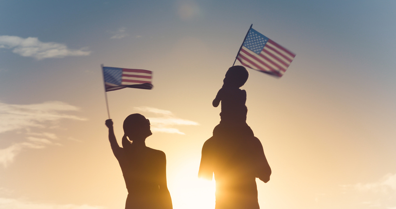 A silhouette of a family with an adult holding an American flag and a child on someone's shoulders also waving a smaller flag, against a sunset sky.