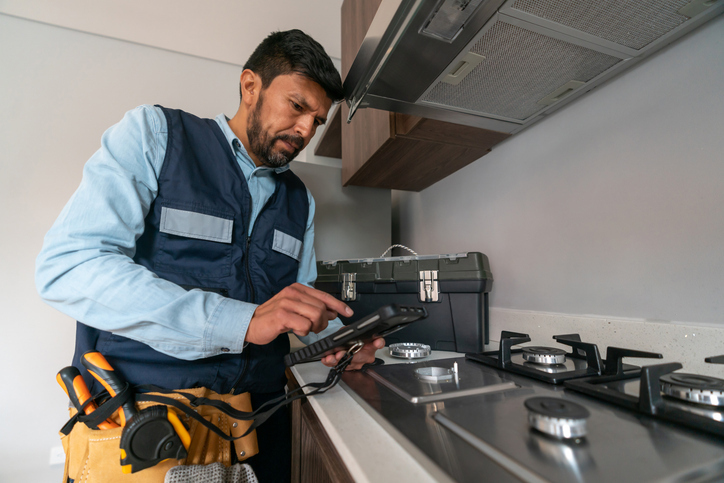 An inspector wearing a tool belt is examining a kitchen appliance with a handheld device.