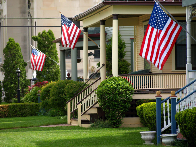 American flags adorn the front porch of a traditional house, creating a patriotic display on a well-maintained property.