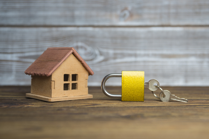 A small wooden model house next to a shiny padlock with keys, set against a rustic wooden backdrop, symbolizing home security or real estate.