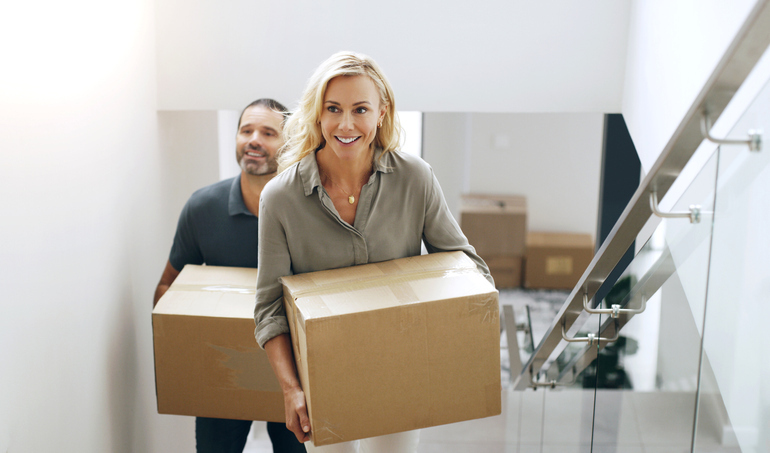 A woman and a man are carrying cardboard boxes up a set of stairs in a bright, modern home, suggesting they are in the process of moving in.