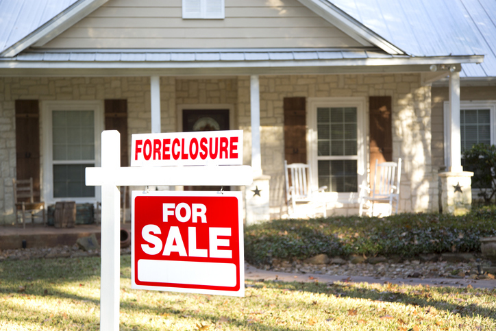 A "For Sale" sign with an additional "Foreclosure" rider is prominently displayed in the front yard of a single-story house, indicating that the property is on the market due to foreclosure.